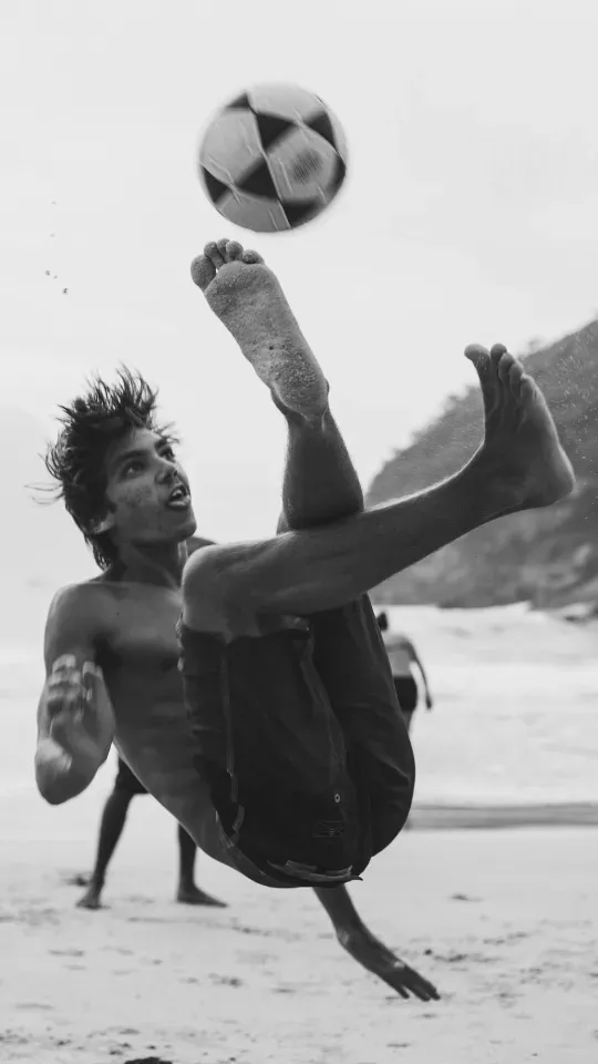 A man is doing a trick with a soccer ball on the beach.