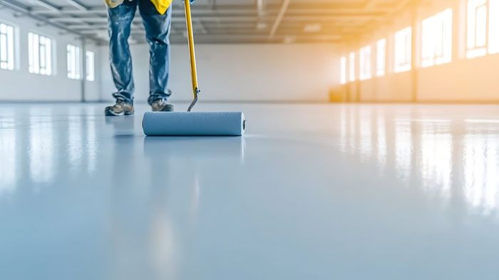 Person applying epoxy floor coating with a roller in a large, empty room.