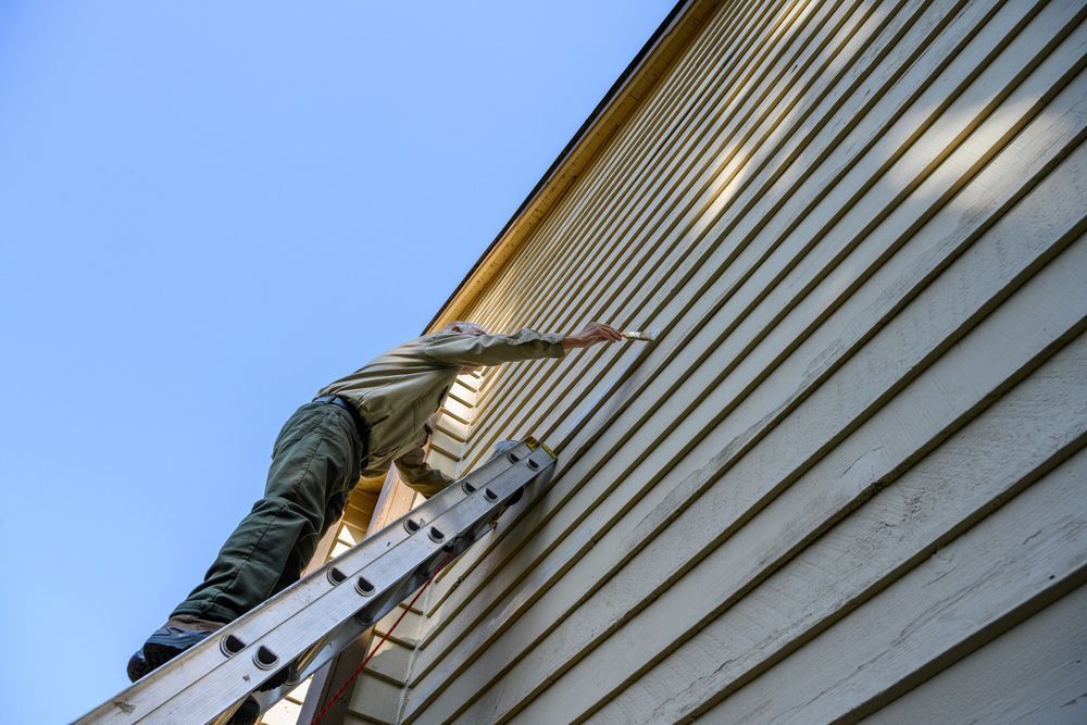 A man is standing on a ladder painting the side of a house.