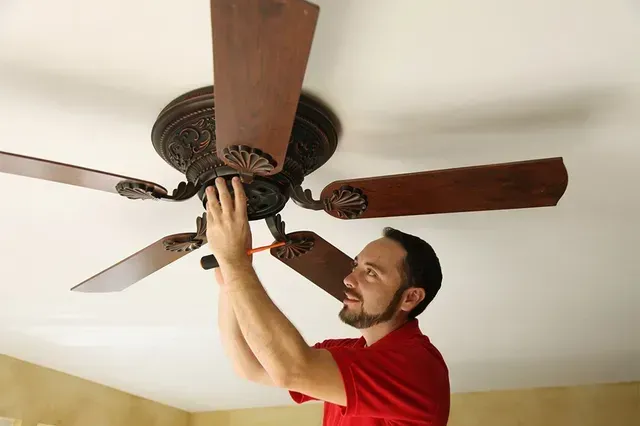 Man adjusting a wooden ceiling fan with a dark ornate mount in a bright room