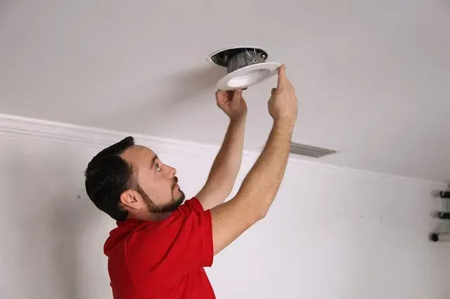 Man in red shirt installing a ceiling light fixture in a white room