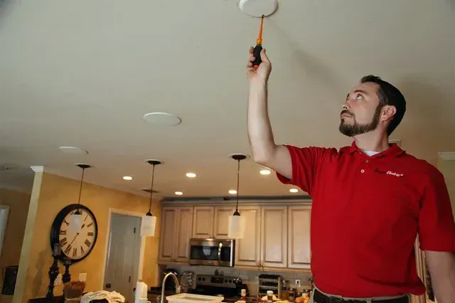 Man in red shirt changing a ceiling light bulb in a kitchen with pendant lights and a wall clock.