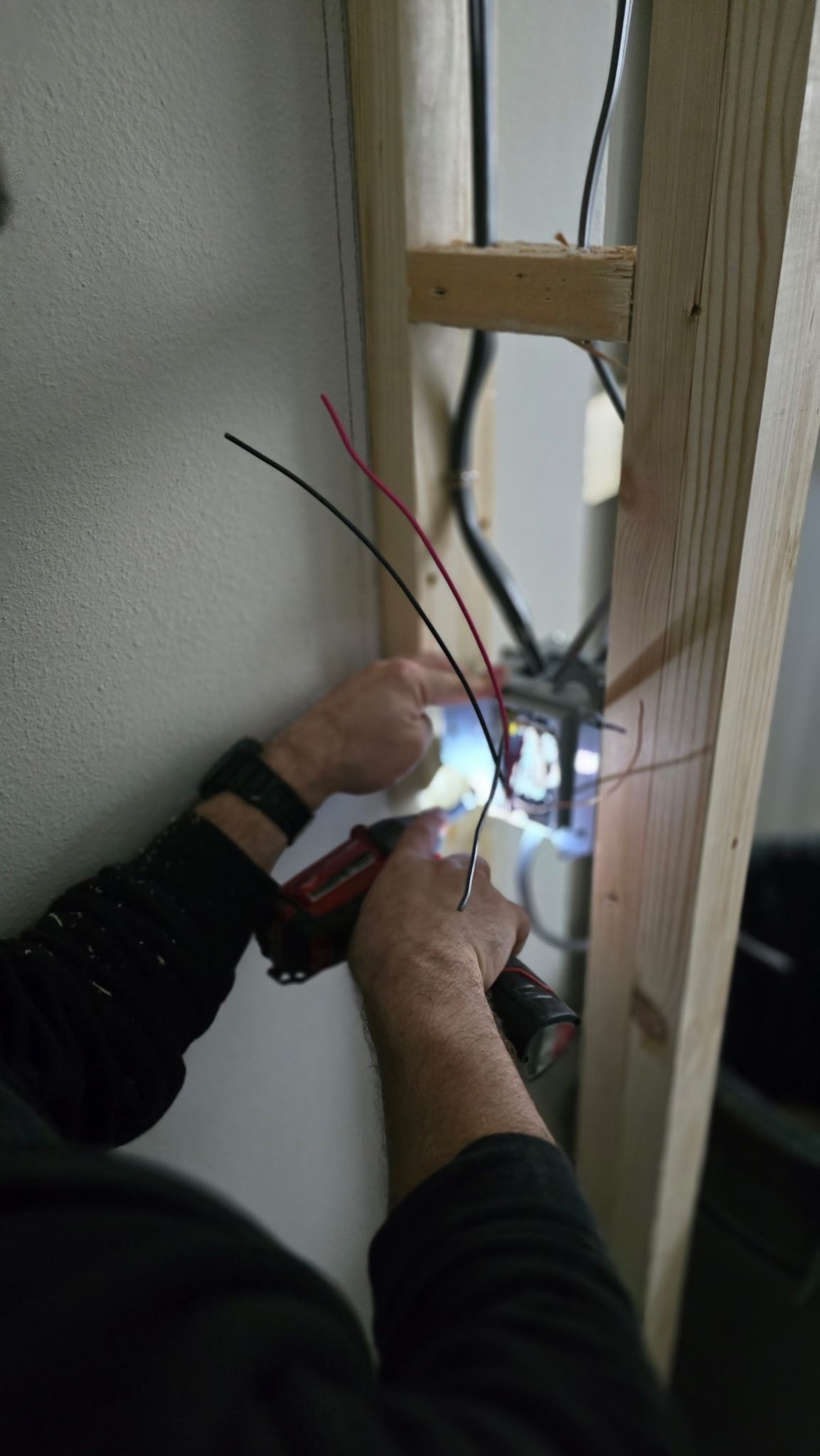Electrician wiring cables in a wall cavity, working with a bright flashlight and exposed studs.