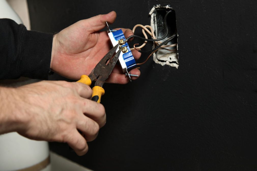 Hands using pliers to cut wires on a blue electrical outlet box in a wall opening