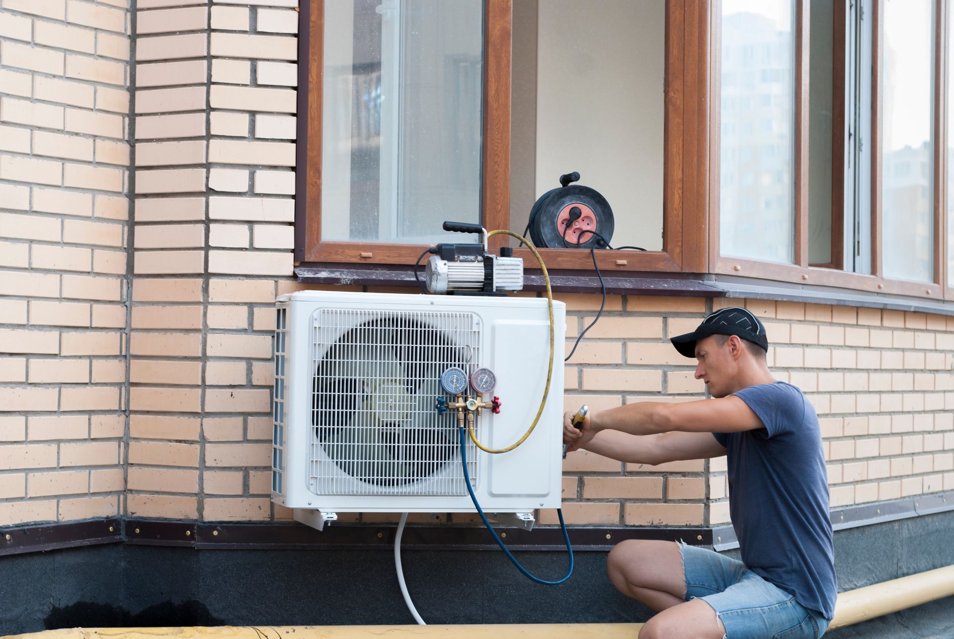 Technician repairing an outdoor air conditioner unit beside a brick house window