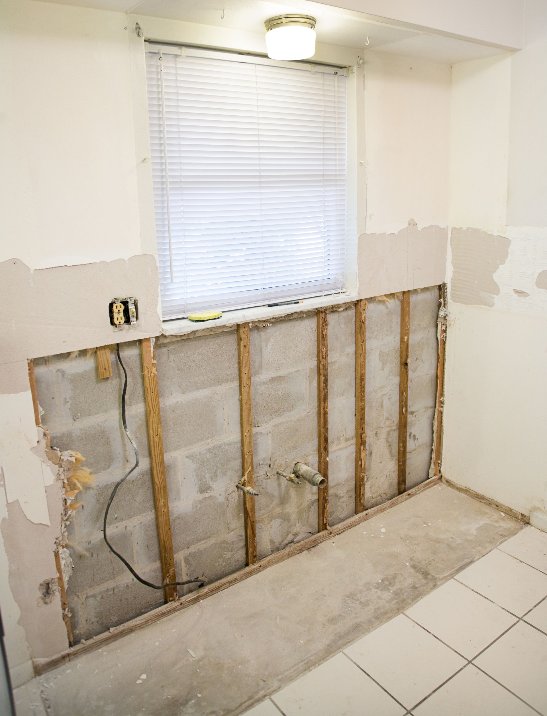 Partially demolished bathroom wall with exposed studs beneath a window and white tile floor