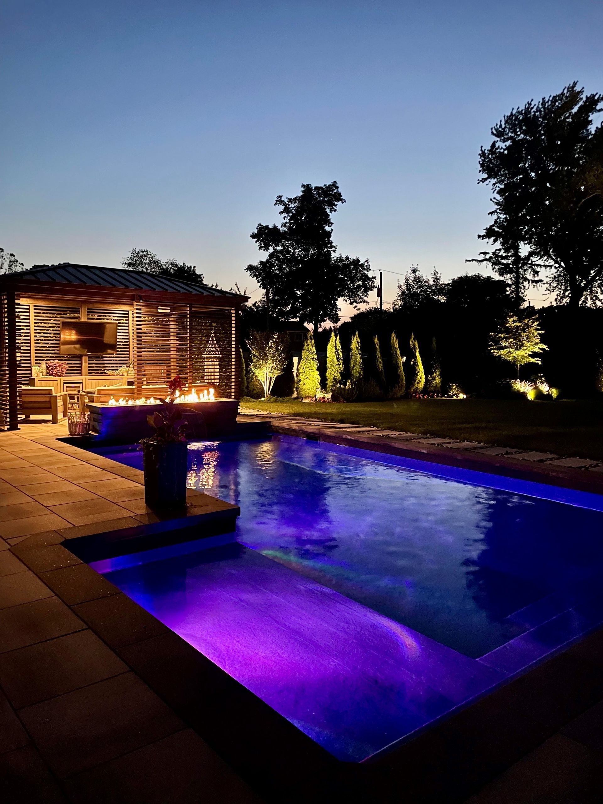 Lit backyard pool at dusk with purple underwater lights and a lit poolside lounge area.