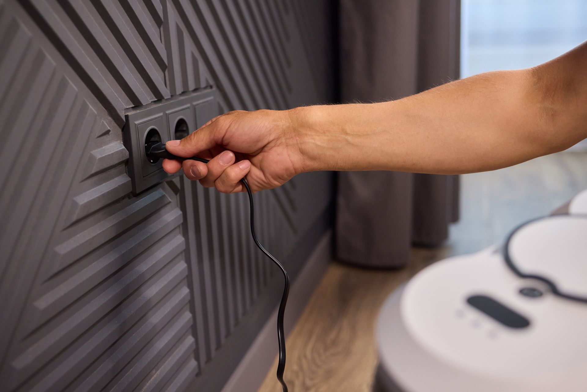 Hand plugging a black cable into a wall outlet beside a white appliance