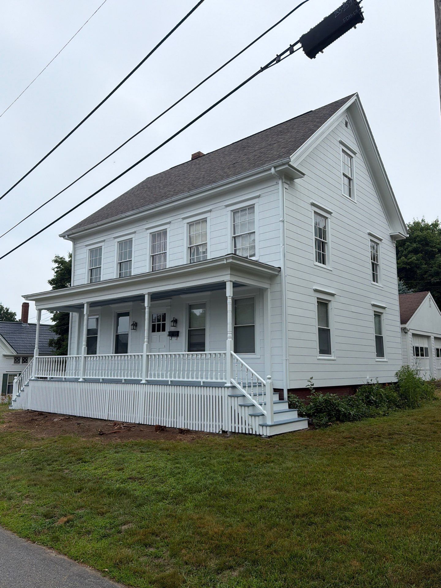White two-story house with a porch and white railing on a grassy lot under overcast skies.