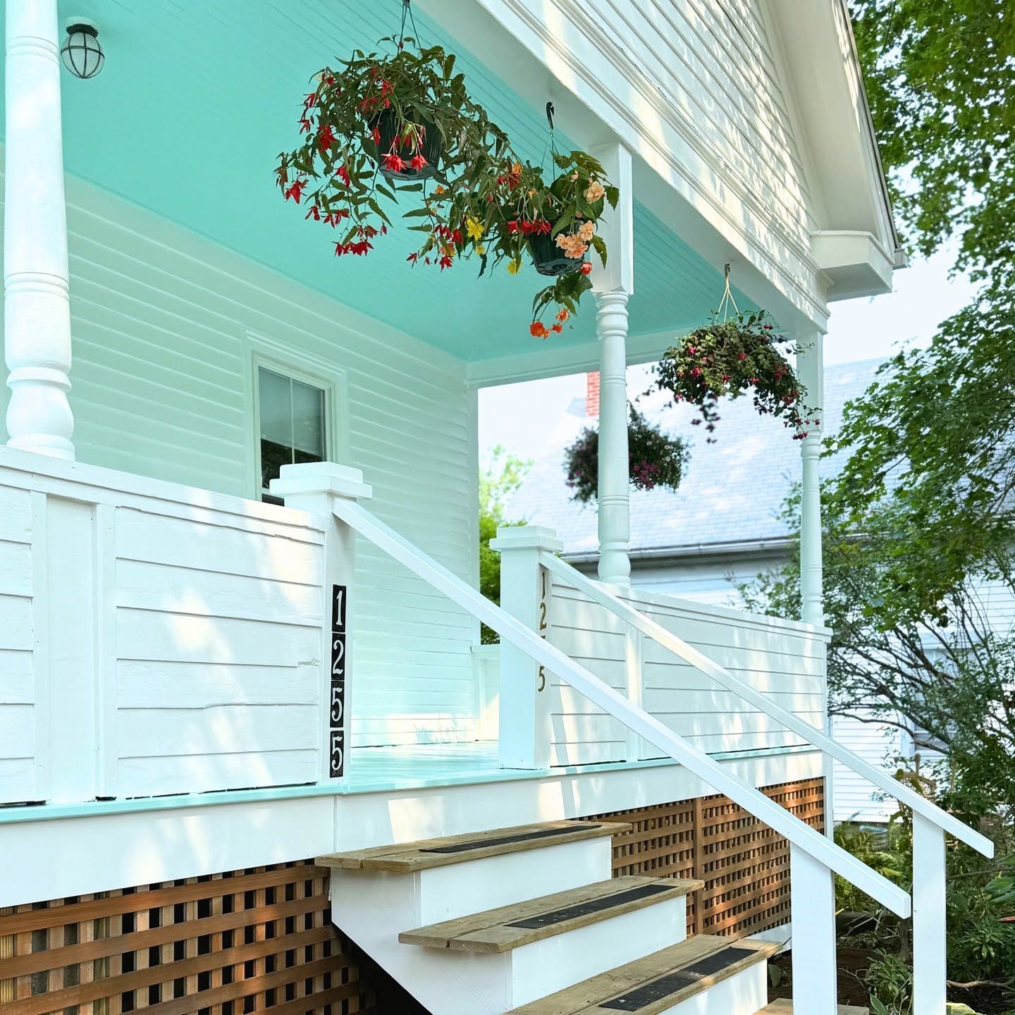 Porch of a white house with hanging flower baskets, white railing, and steps above a wooden lattice skirt