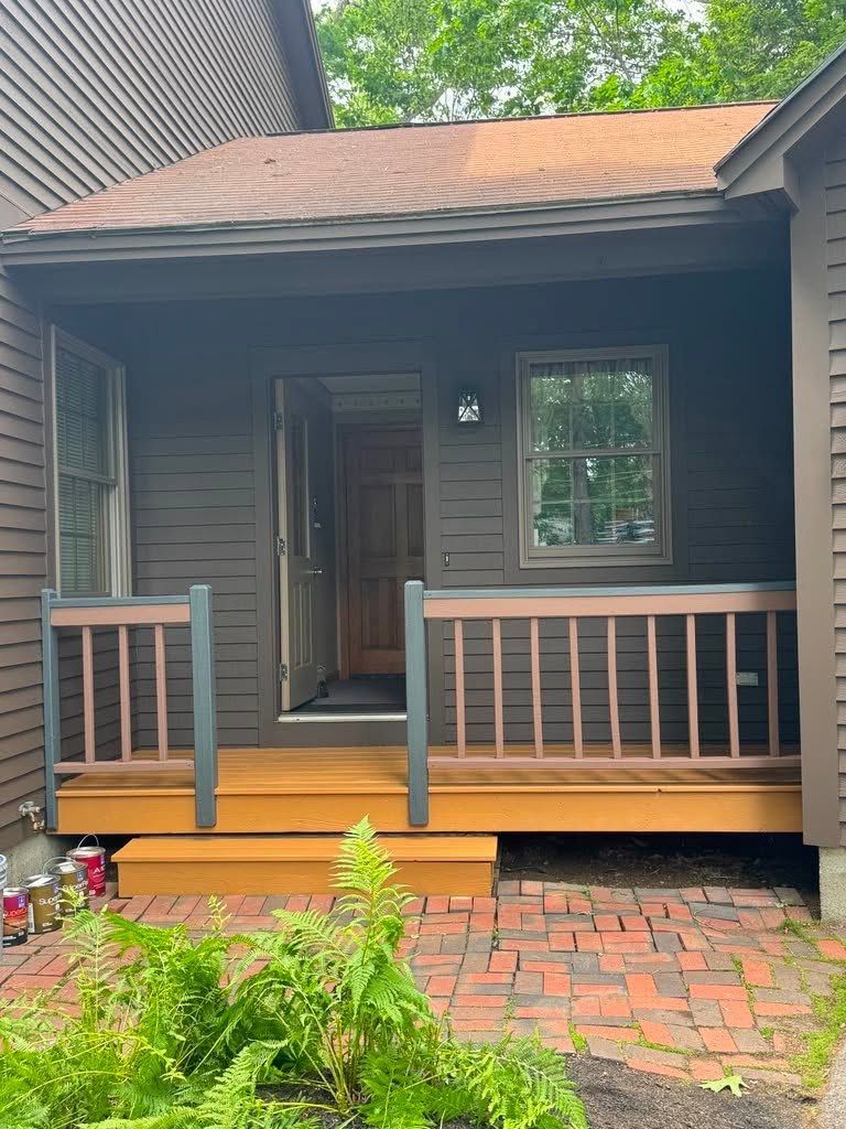Small wooden porch on a dark house with gray railings, brick path, and front door under a shingled roof