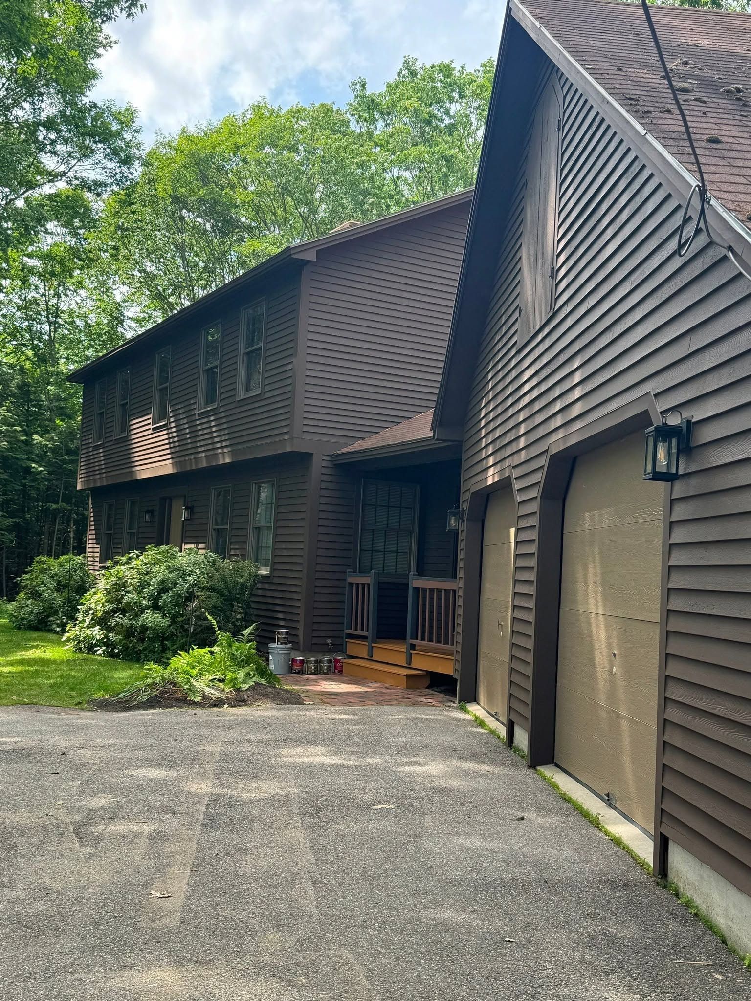 Wood-sided house with steep roof, garage door, and a gravel driveway surrounded by trees
