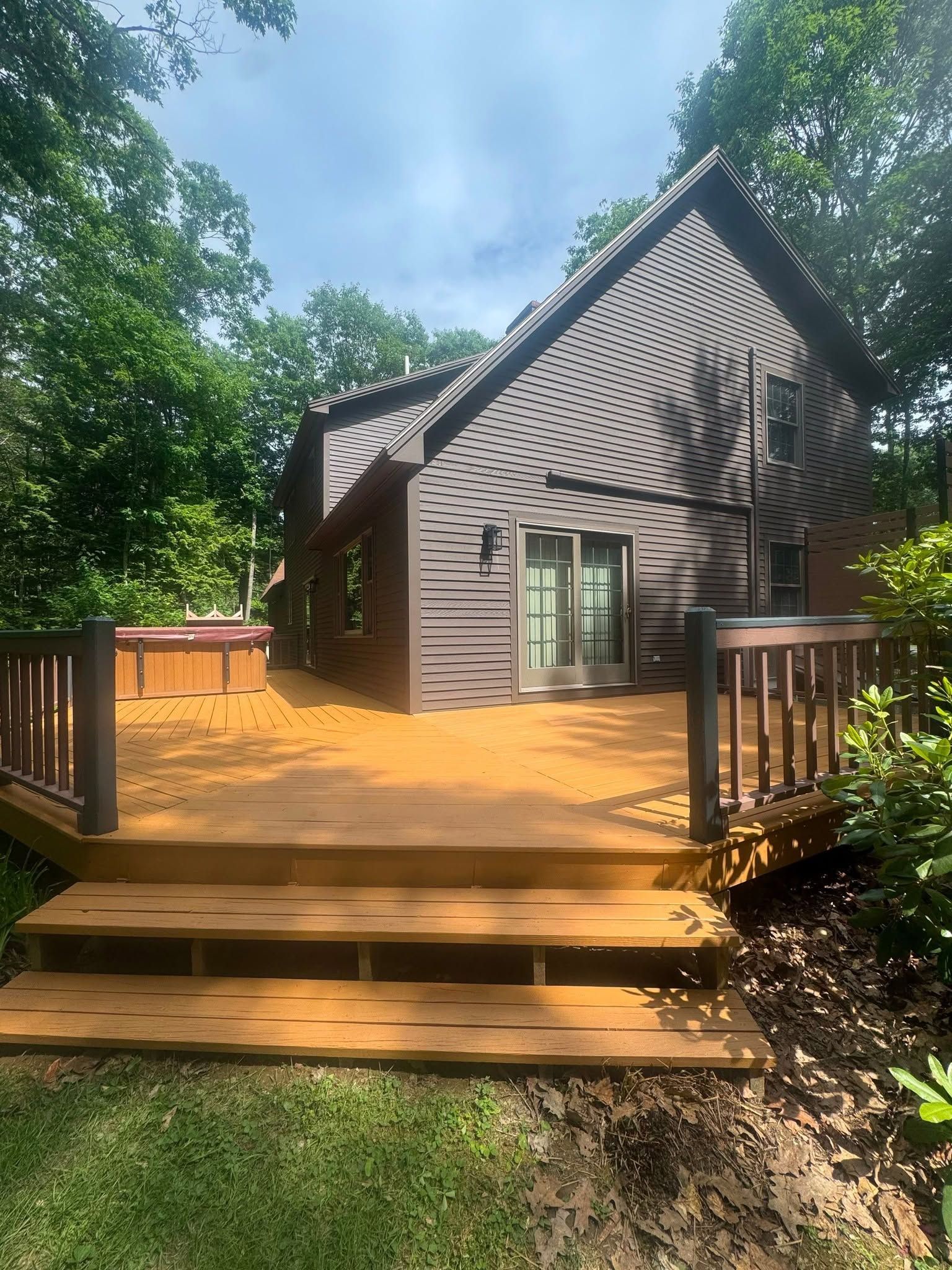 Wooden steps lead to a gray house with a sunlit deck surrounded by trees.