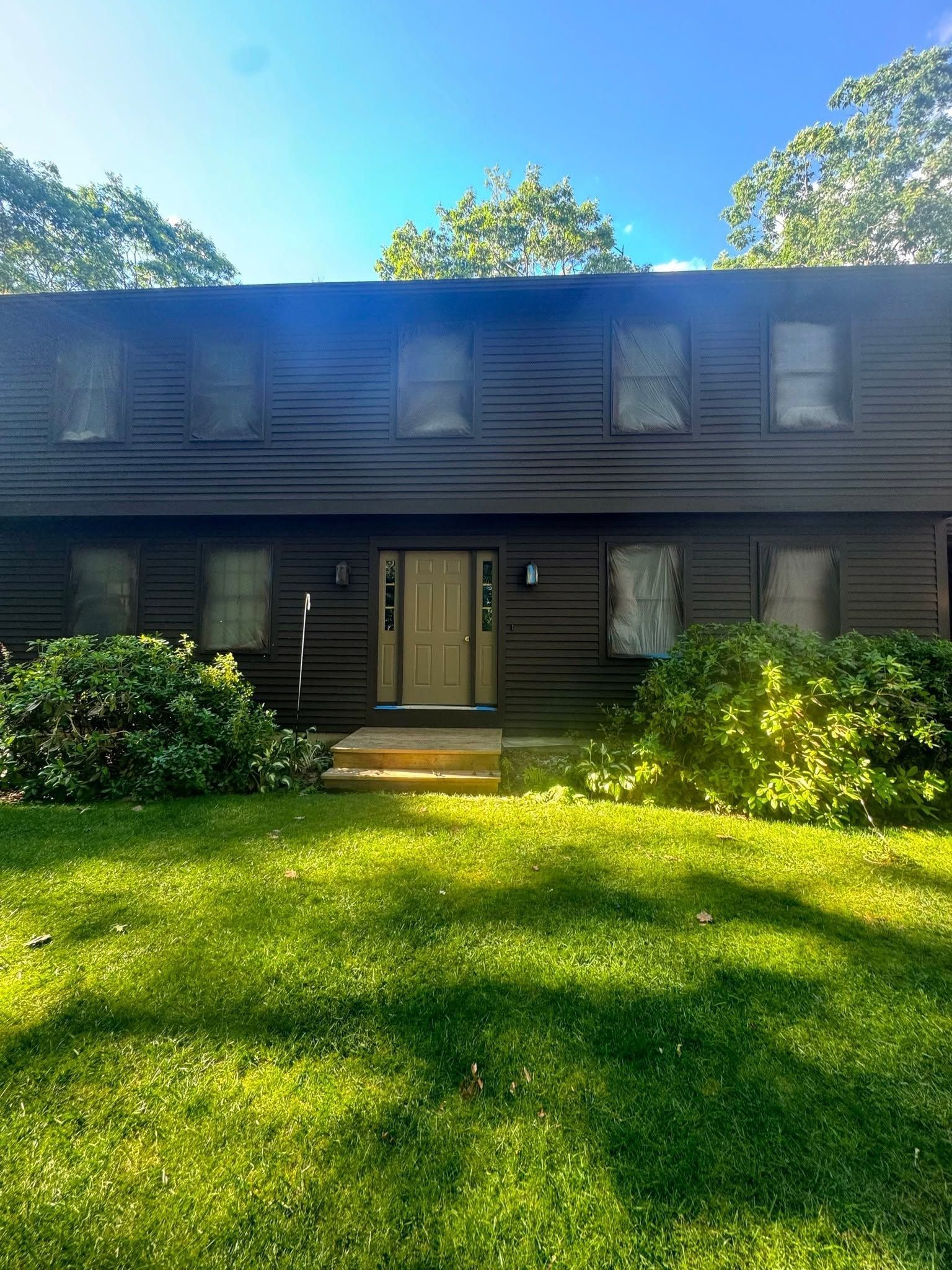 Two-story dark house with white shutters and a front lawn in bright sunlight.