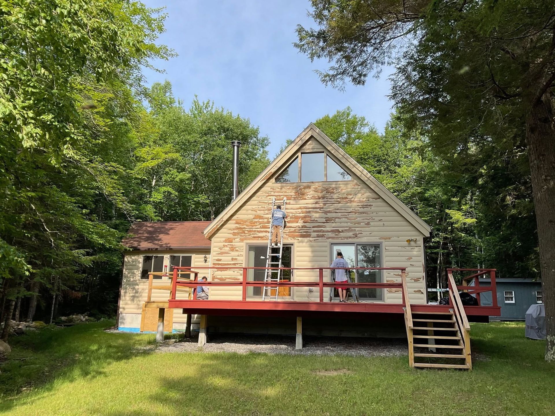 Small beige cabin with red deck and stairs in a wooded yard under a clear sky