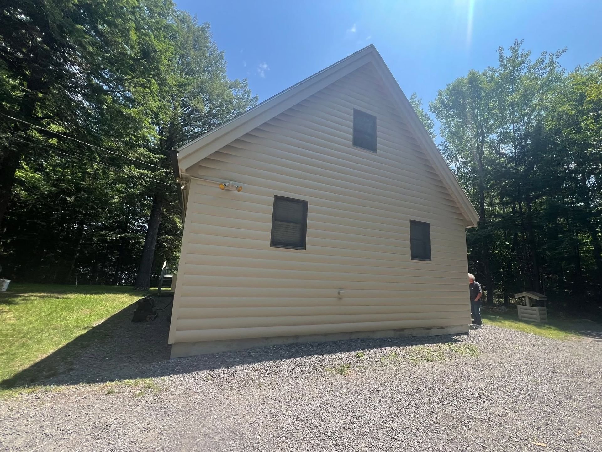 Small beige cabin with dark windows beside a gravel driveway, surrounded by trees in bright sunlight