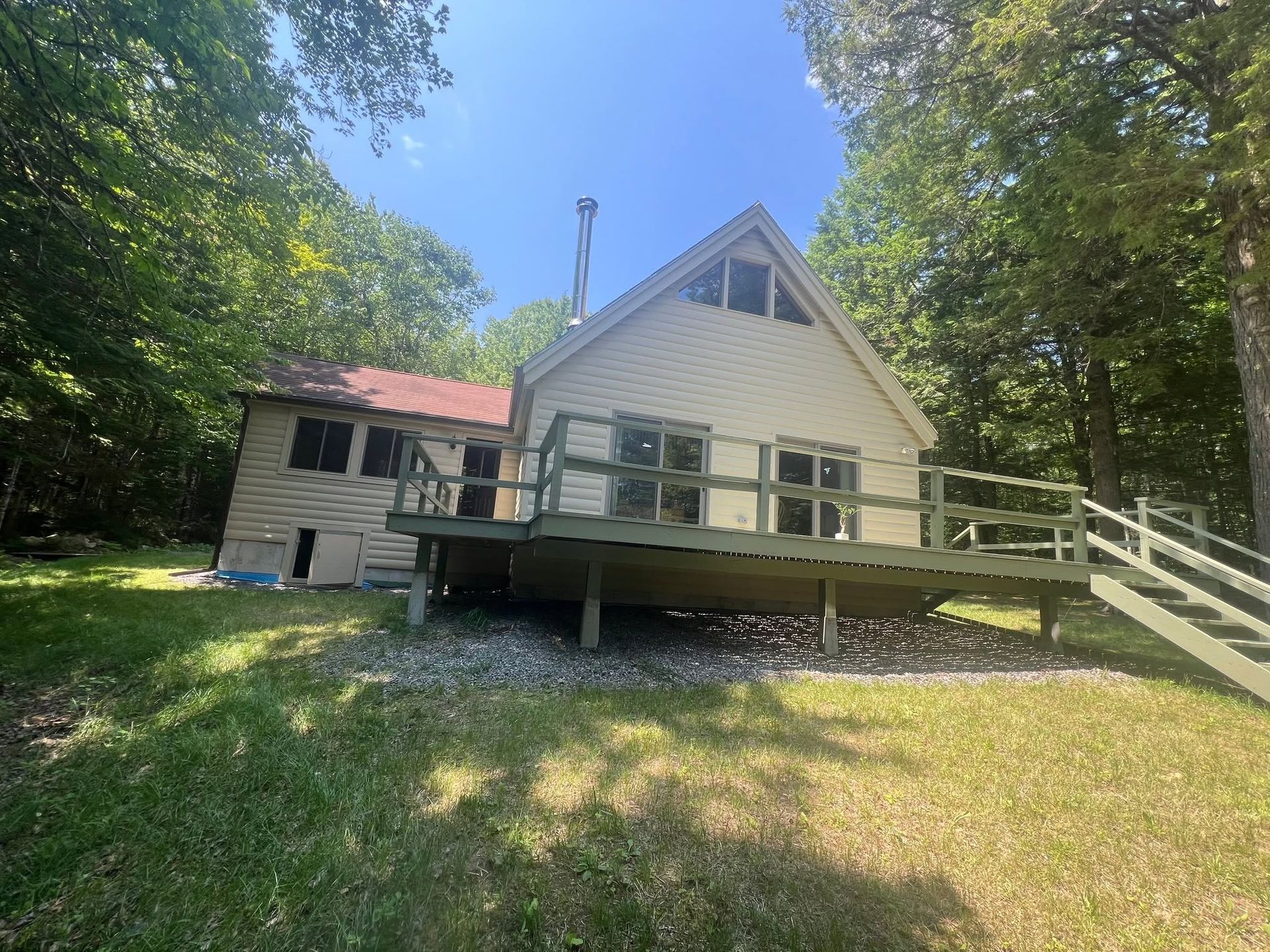 White A-frame cabin with green deck and ramp, surrounded by trees on a sunny day