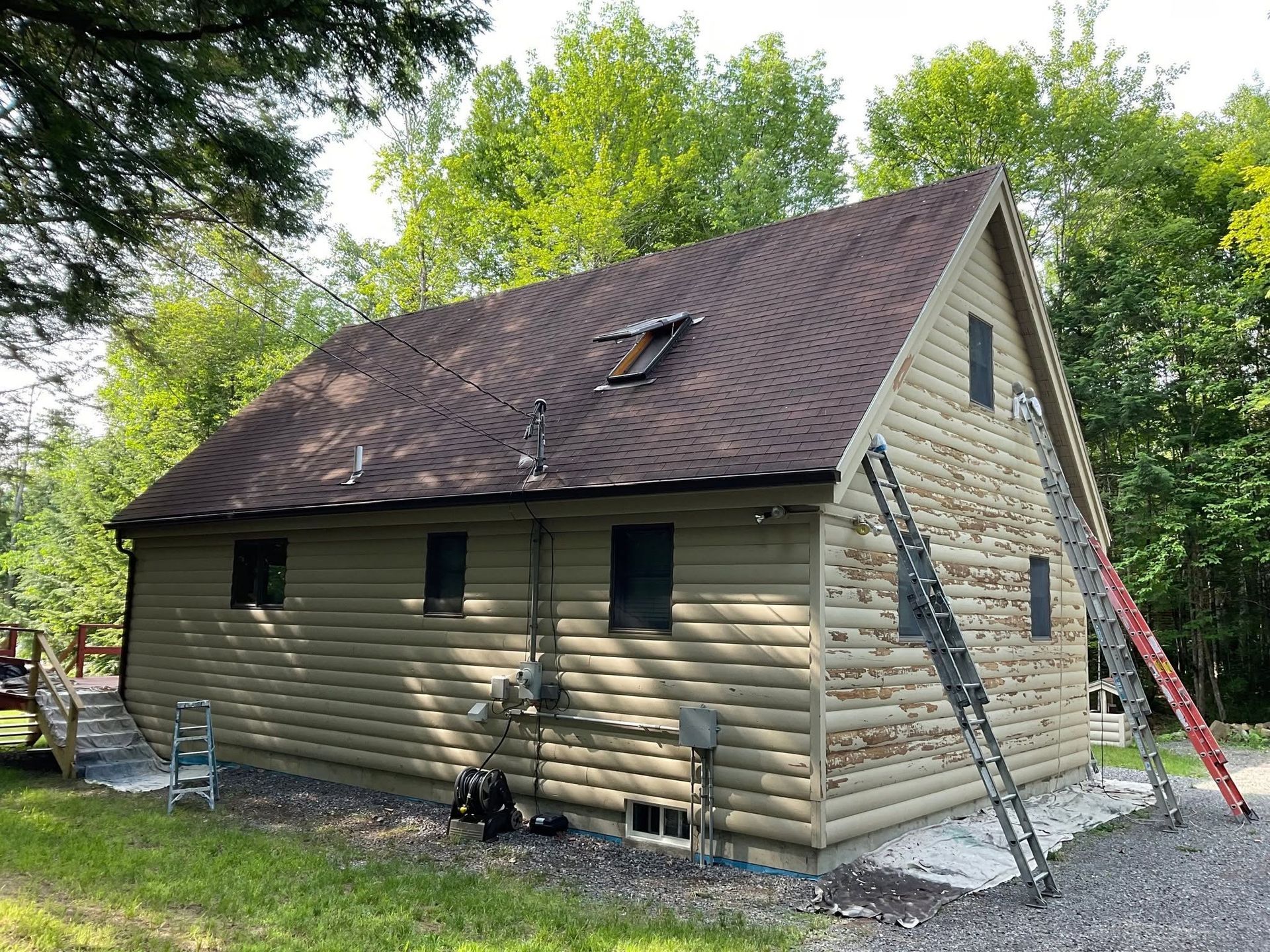 Small stone house with a steep brown roof, ladders, and workers’ tools in a wooded yard.