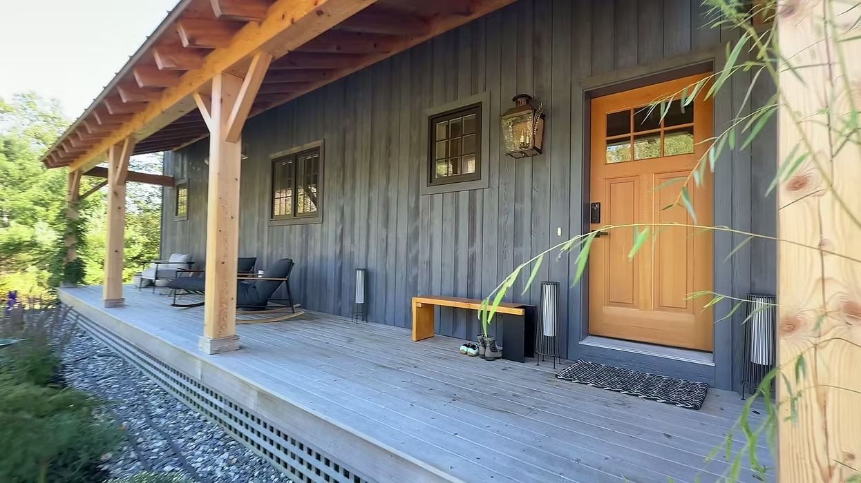 Rustic cabin porch with wooden posts, a bench, and a brown door beside a dark exterior wall.