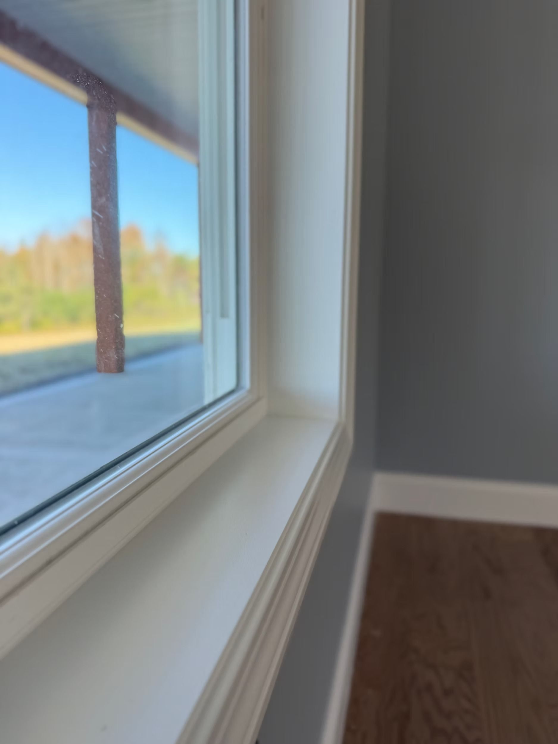 Close-up of a white window frame overlooking a blue sky and trees, with a gray wall and wood floor inside
