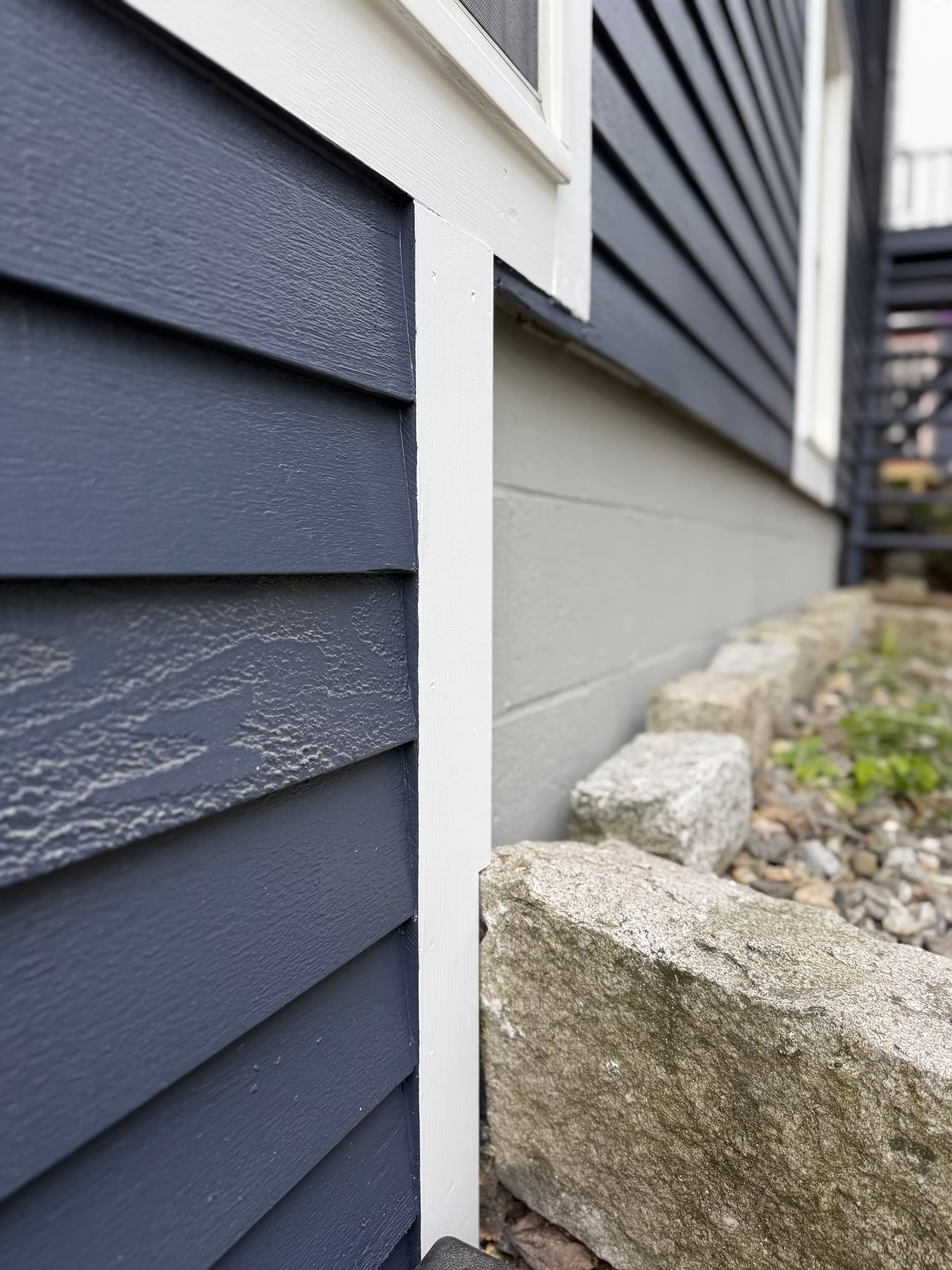 Close-up of blue siding, white trim, and a gray foundation beside a gravel bed and stone edging