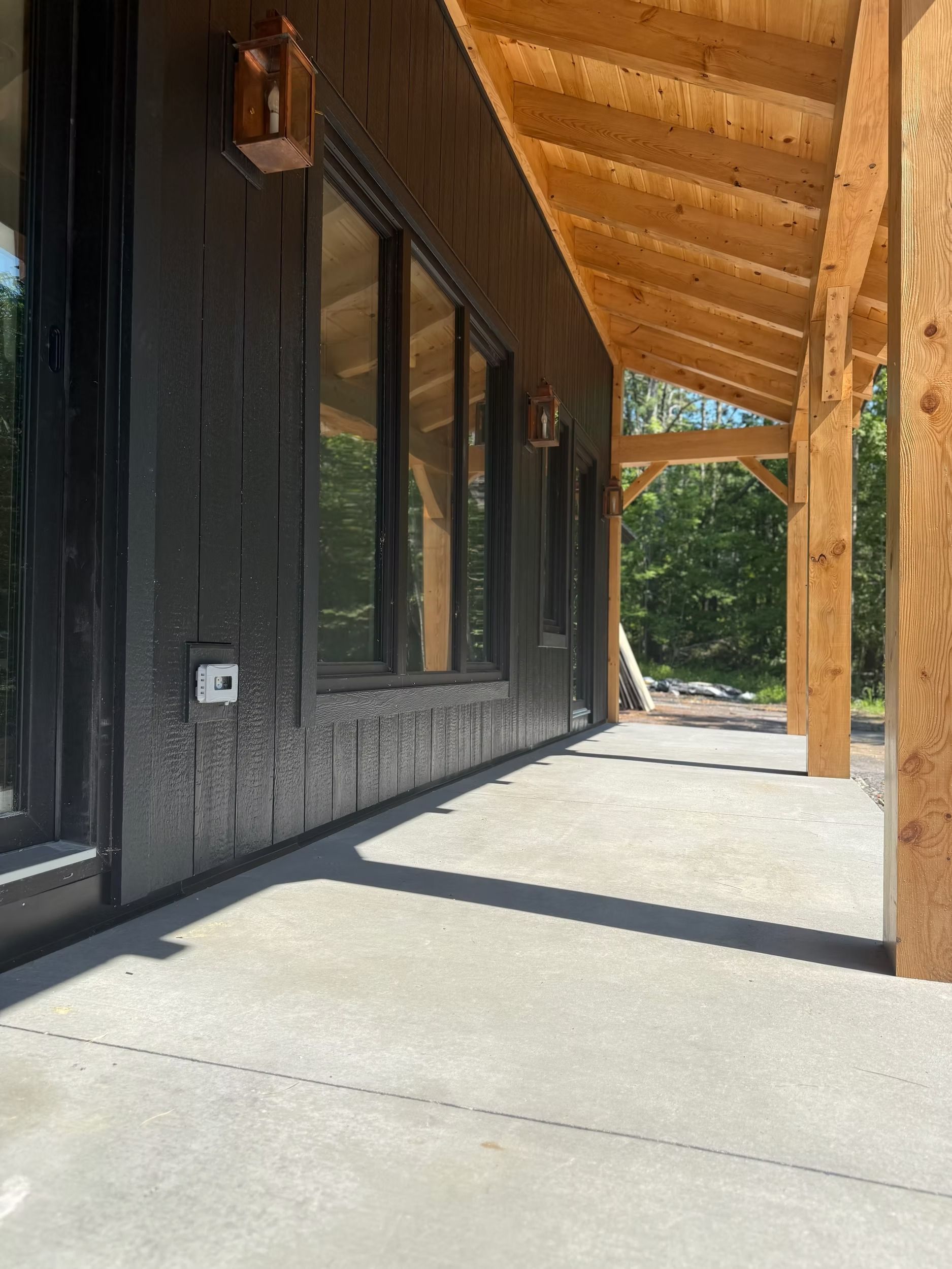 Covered wooden porch with black siding, concrete floor, and timber beams leading to trees outside