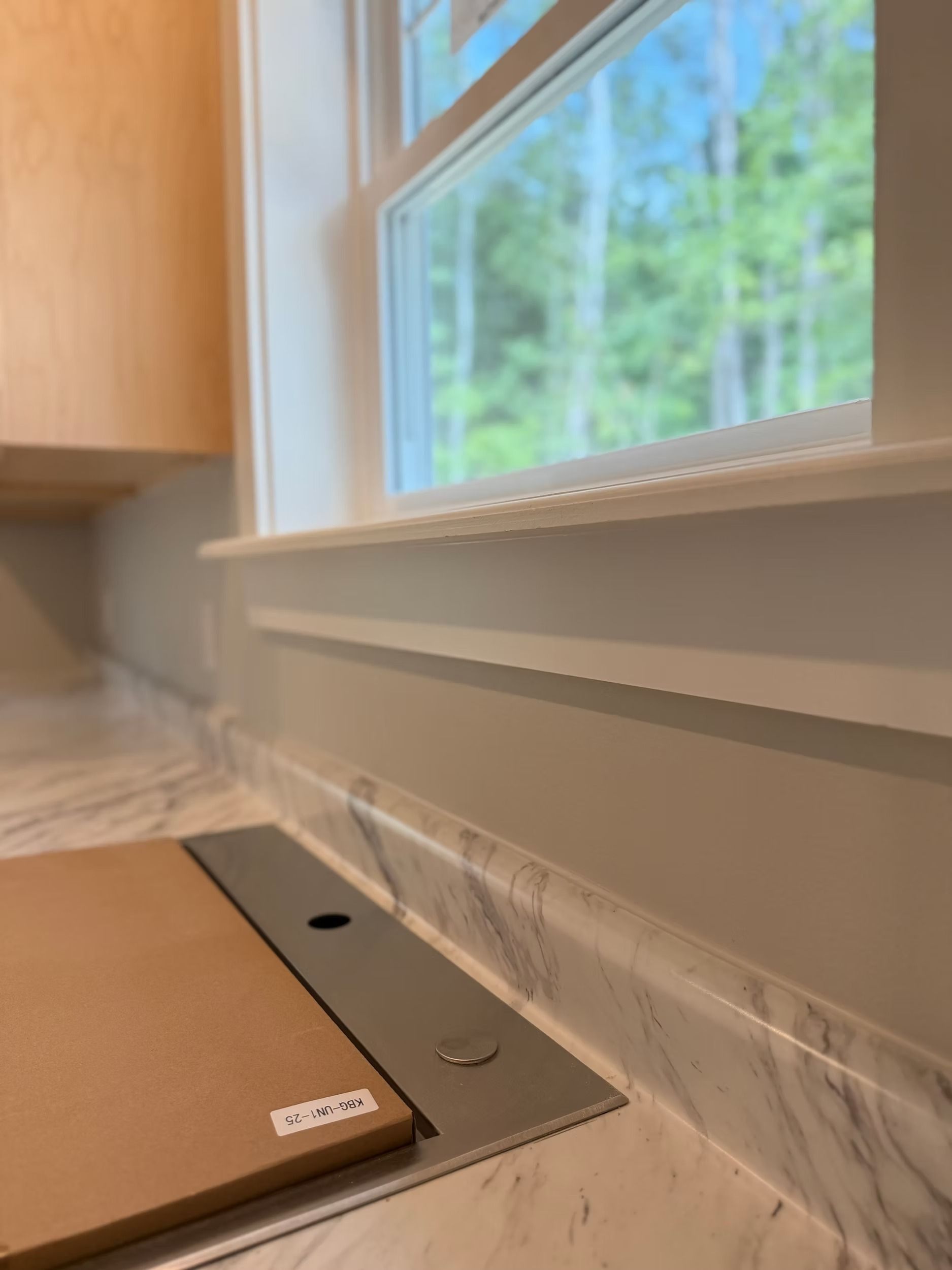 Floor-level view of a room corner with a window, baseboard, and a metal floor plate near a cardboard box.