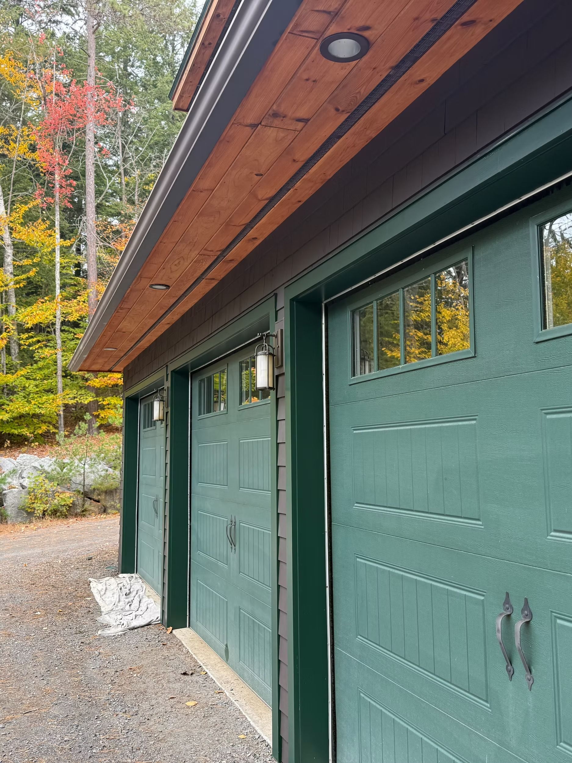 Green garage doors under a wooden overhang beside a gravel driveway and autumn trees