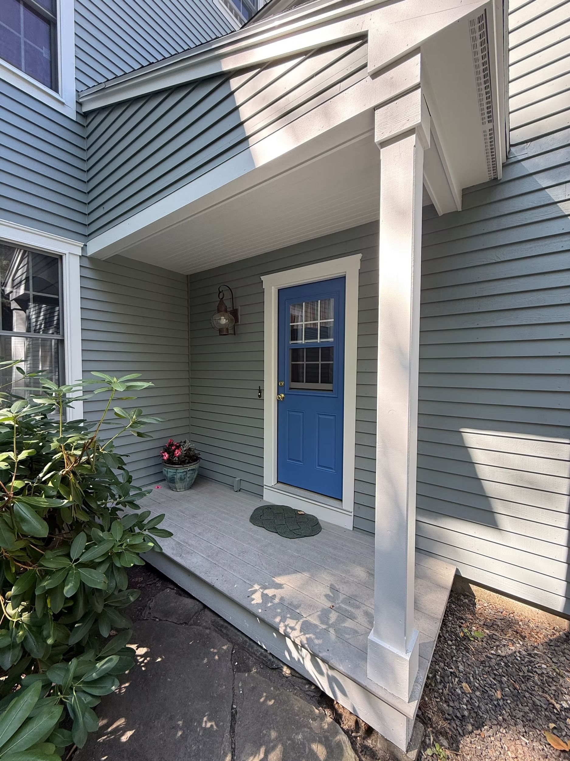 Blue front door under a covered porch on a gray house, with white trim and potted plants.