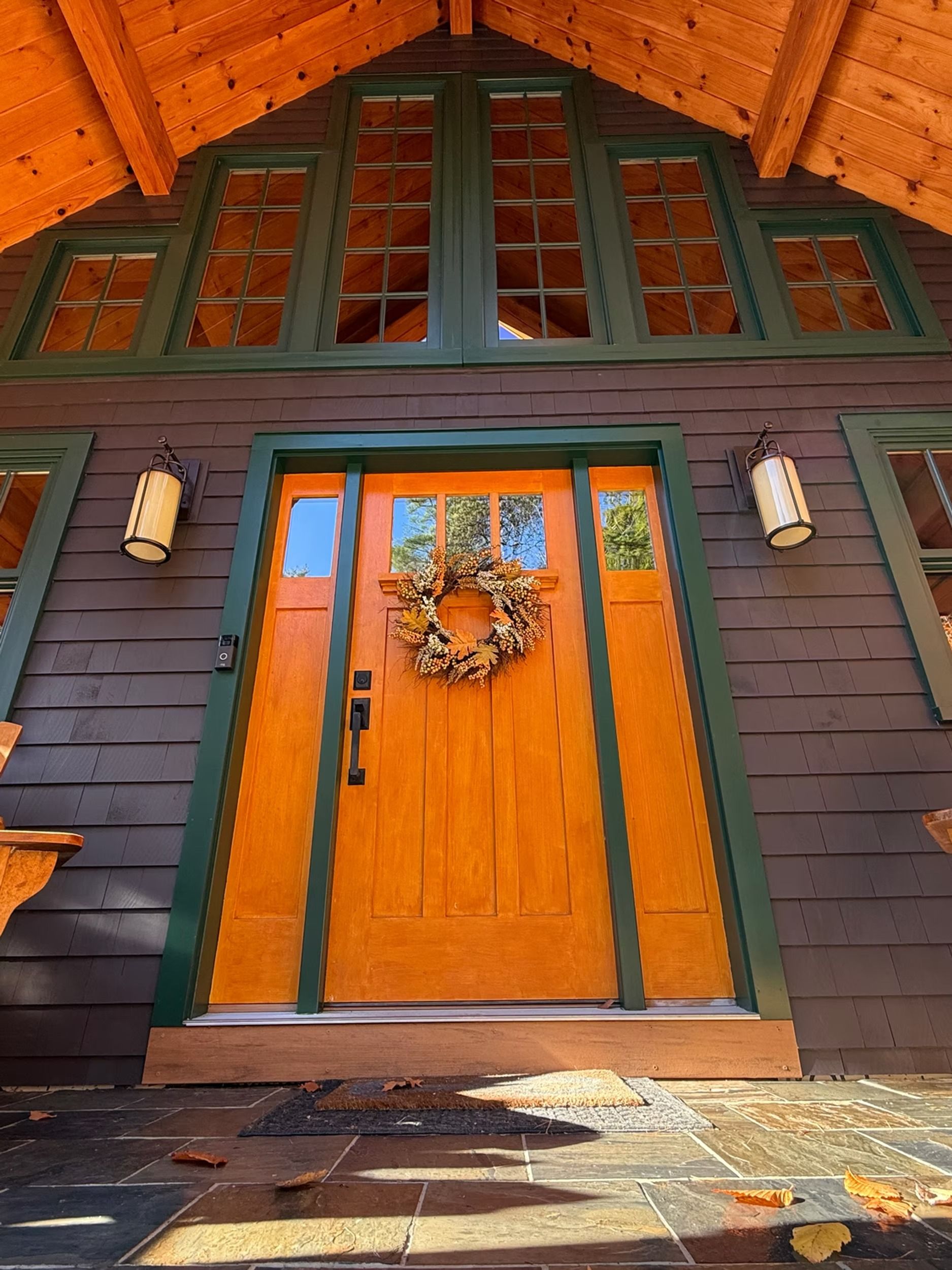 Orange front door with wreath in a rustic dark green cabin facade, under a peaked wood roof.