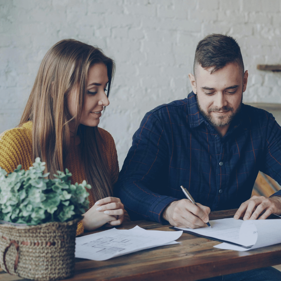 Two people smiling while signing documents at a table in a bright office