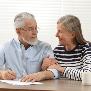 Two older adults smiling at a table while reviewing paperwork with a pen and coffee mug nearby