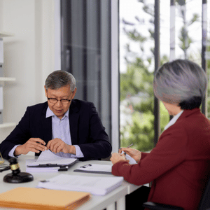 Two people in an office meeting, one writing on papers while the other listens across a desk.