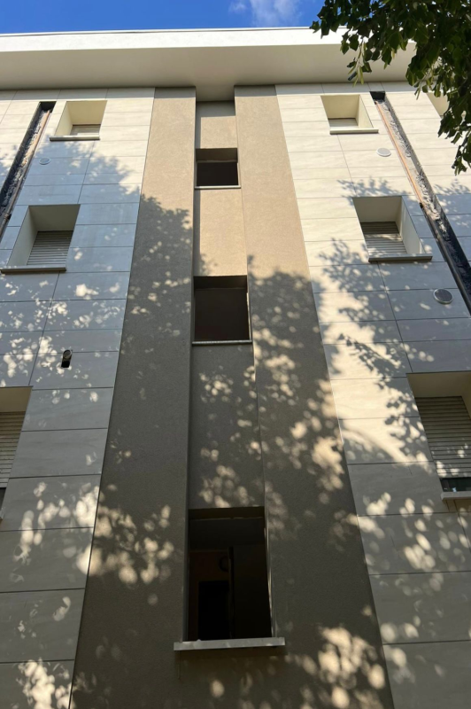Facciata di un alto edificio residenziale beige con balconi e ombre degli alberi.