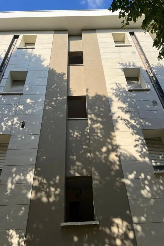 Facciata di un alto palazzo residenziale beige con finestre e ombre di alberi illuminate dal sole.