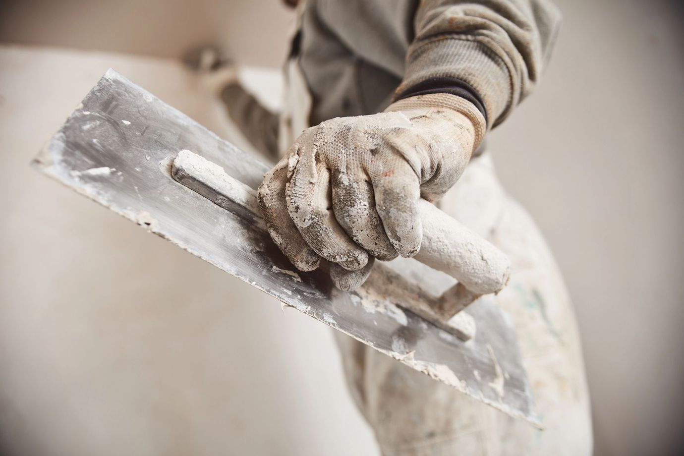 Mano guantata che impugna una cazzuola da intonaco ricoperta di gesso bagnato, applicandolo a una parete.