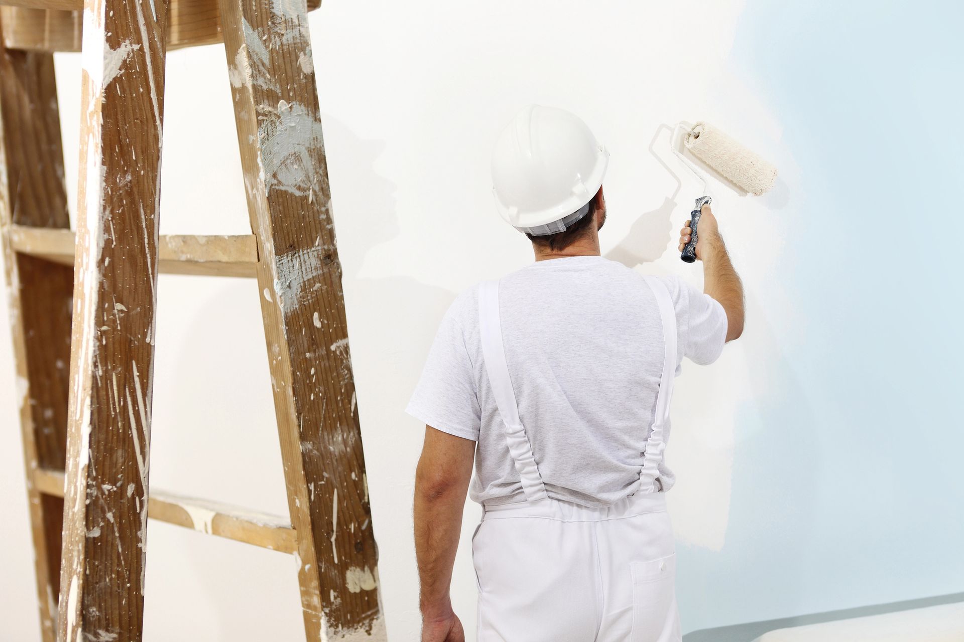 Un imbianchino in tuta bianca e casco protettivo dipinge un muro bianco accanto a una scala di legno.