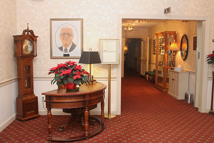 Elegant hallway with grandfather clock, round table of red flowers, and framed portrait.