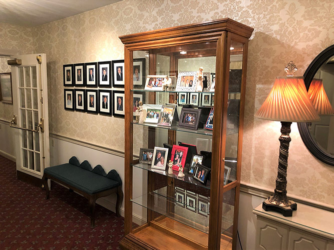 Hallway with framed photos, a glass display cabinet, bench, and wall sconce lamp on patterned wallpaper.