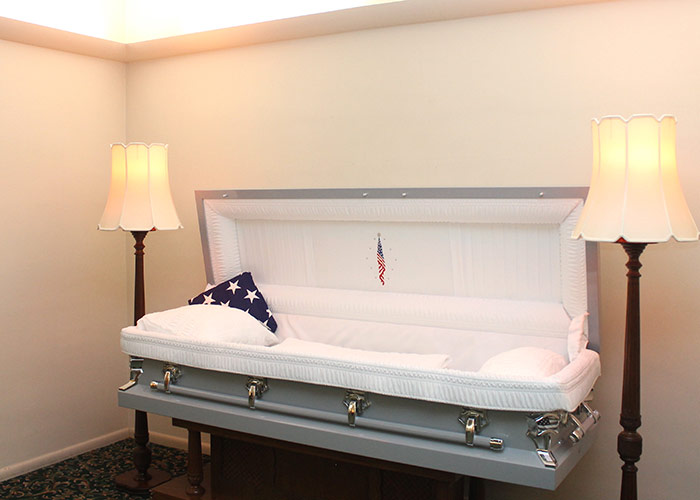 White coffin with American flag pillow on stands, lit by two lamps in a viewing room