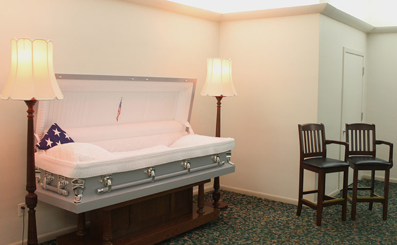White casket in a funeral home room with lamps and chairs beside it