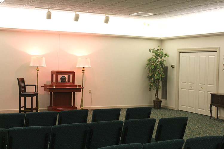 Empty chapel with rows of seats, altar table, two lamps, and closed double doors.