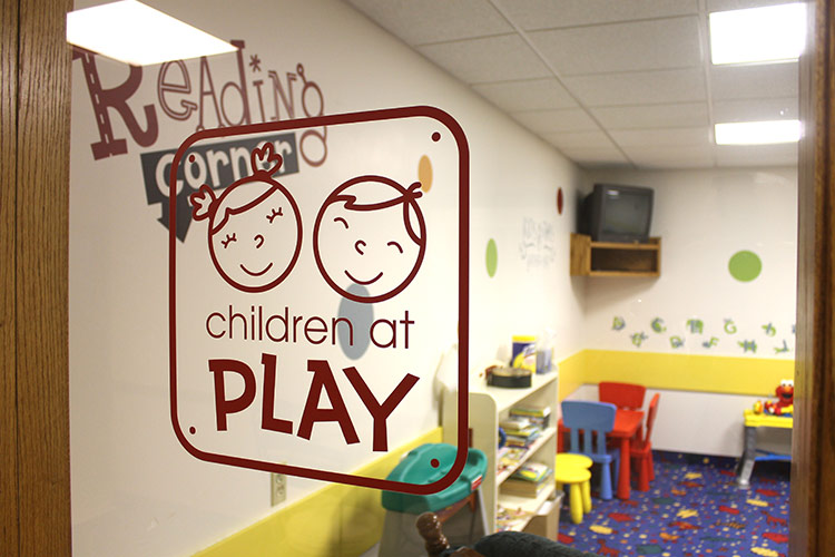 Play corner in a children’s room with “children at PLAY” sign, small tables, chairs, and bright wall decor.