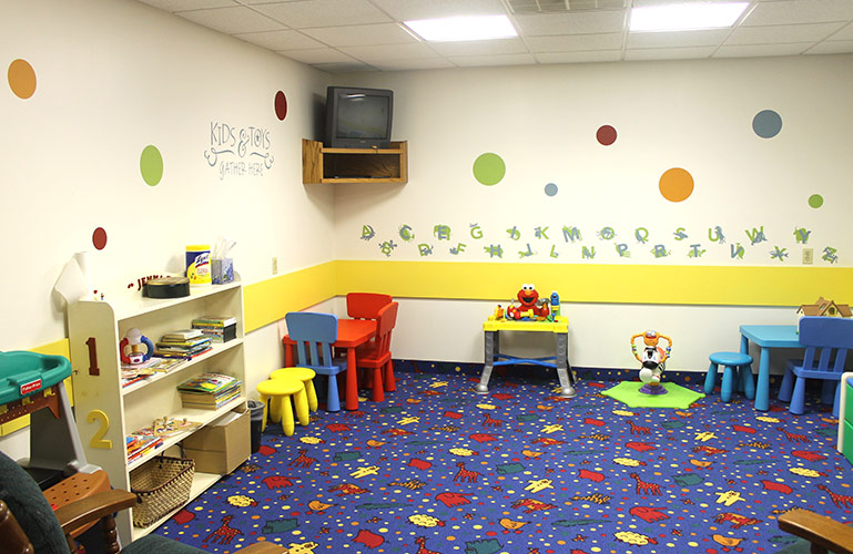 Colorful preschool playroom with small tables, chairs, shelves, and toys on a patterned blue carpet