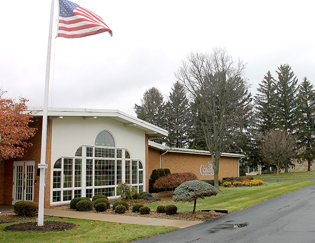 American flag flying beside a building with large arched windows, surrounded by trees and a wet driveway