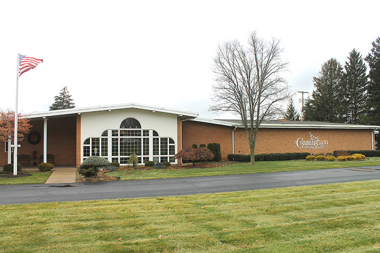 Low brick building with arched windows and an American flag in front, set beside a lawn.