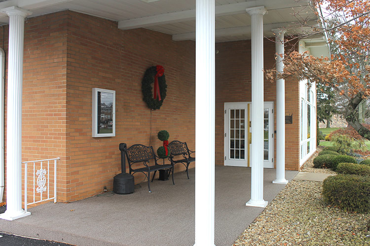 Brick porch with white columns, bench, wreath, and trimmed shrubs along a walkway