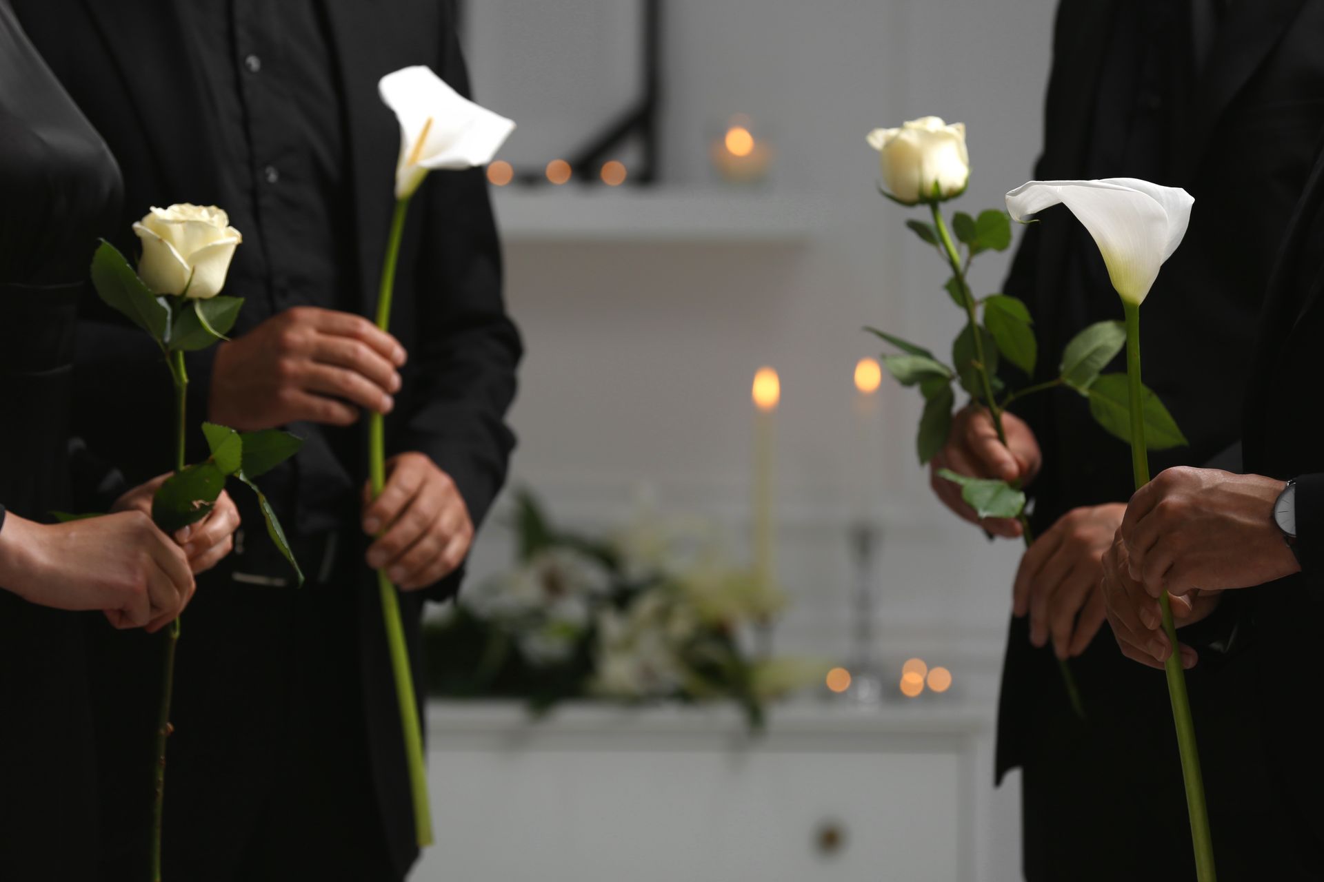 A person in a black blazer holds a single white rose with green leaves against a dark background.