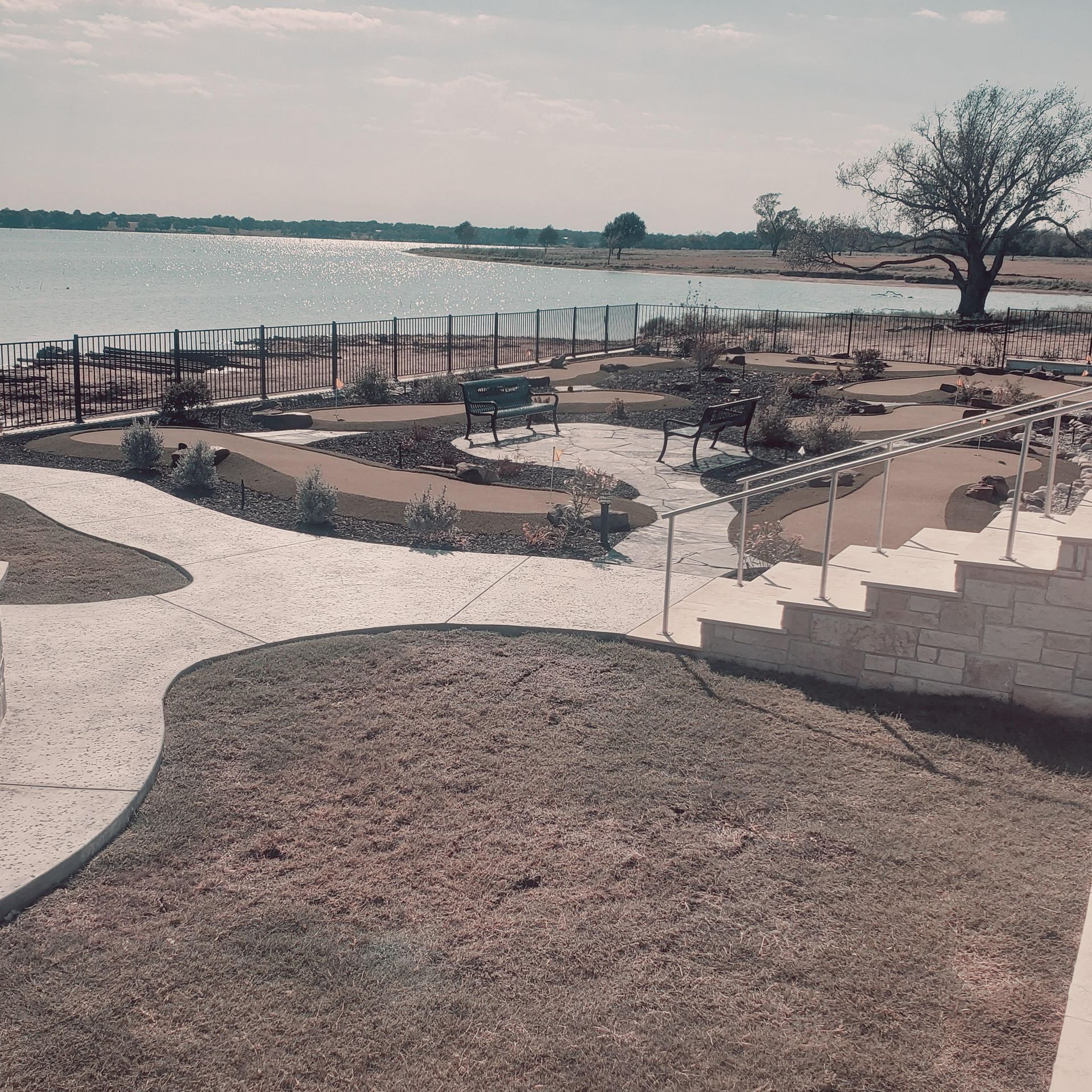 Lakefront garden with winding stone paths, benches, and a lone tree beside calm water.
