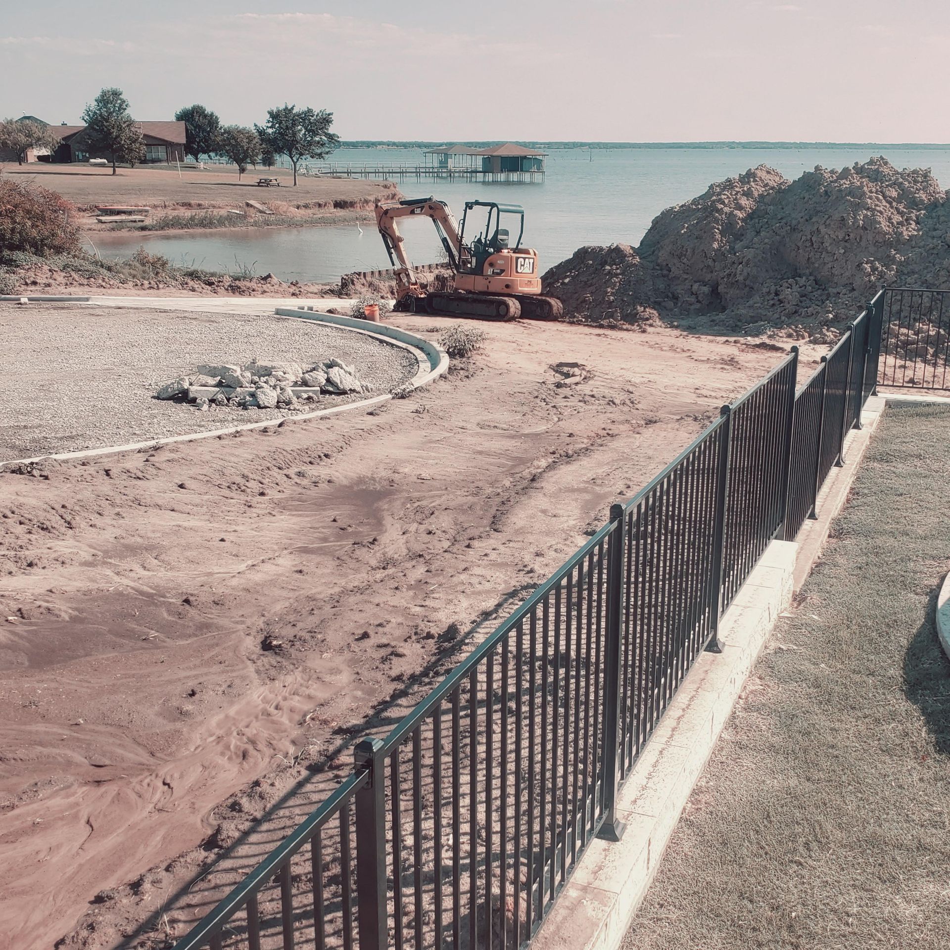Construction excavator on a sandy lakeside path beside a black metal fence and calm blue water.